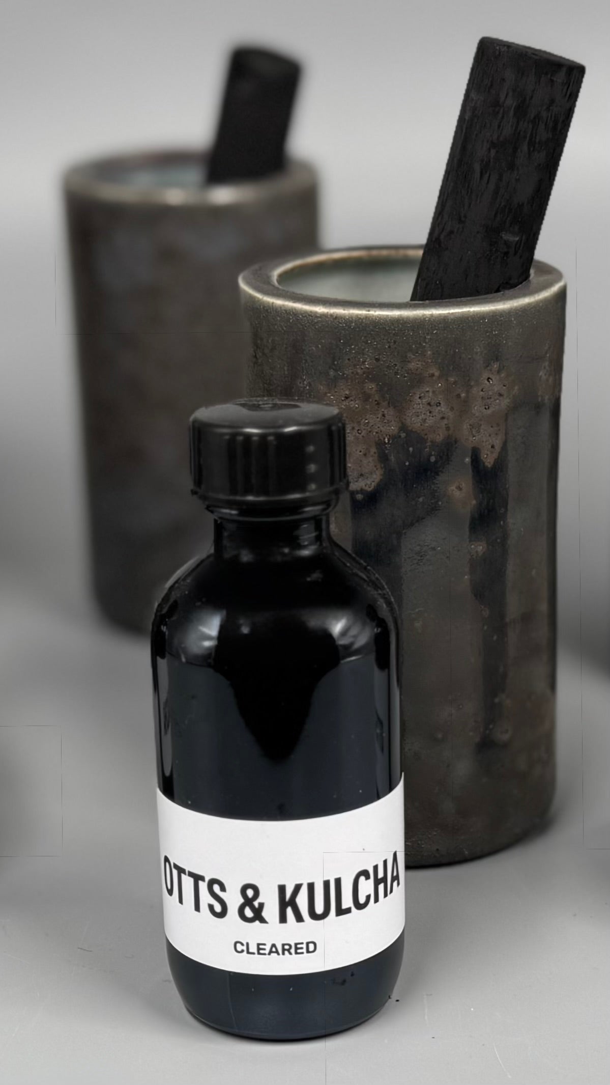 Bottle labeled 'OTTS & KULCHA' with two cylindrical containers in the background on a gray surface.