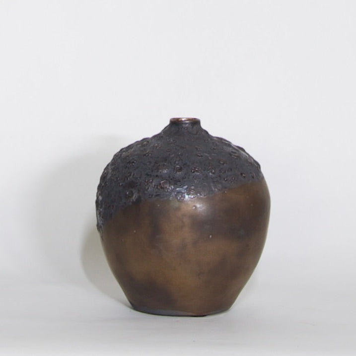 A handmade ceramic vase with a rounded, bulbous shape, featuring a dark, textured, crackled glaze on the upper half and a smooth, metallic bronze-brown glaze on the lower half, set against a plain light gray background.
