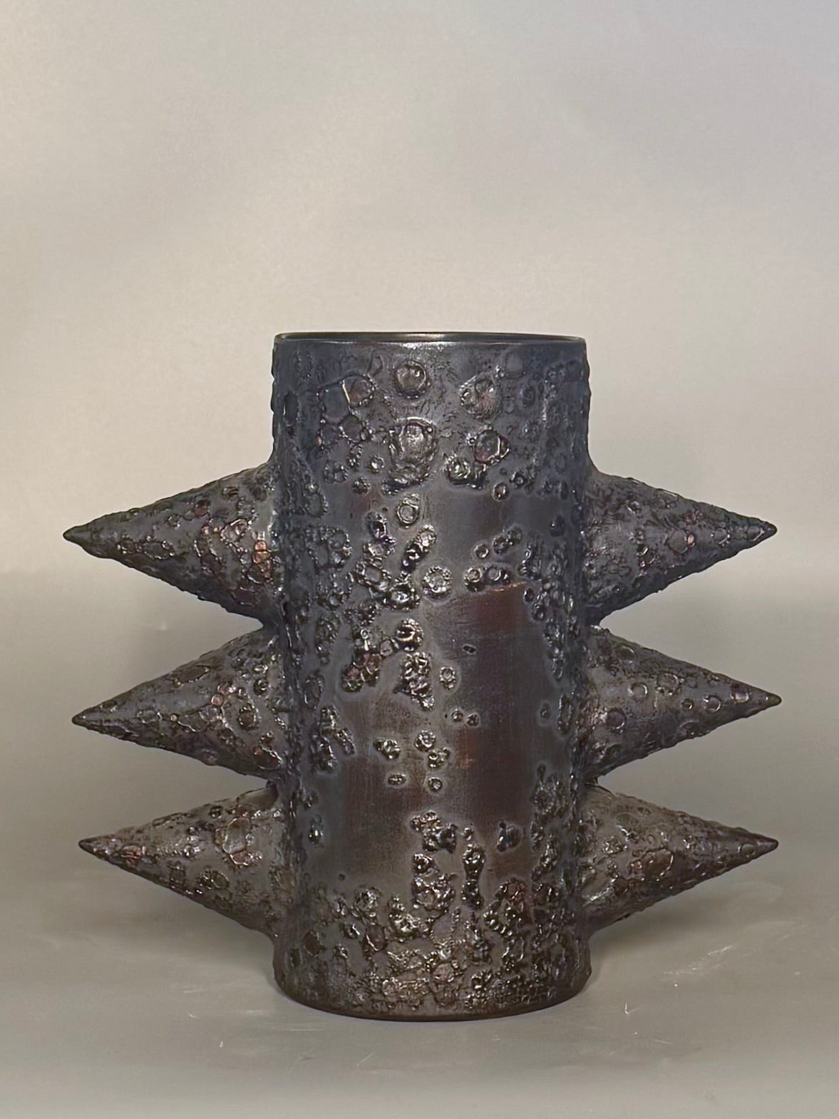 A dark metallic cylindrical sculpture or vase with three rows of sharp conical spikes and a heavily textured, bumpy surface, photographed against a plain light gray background.