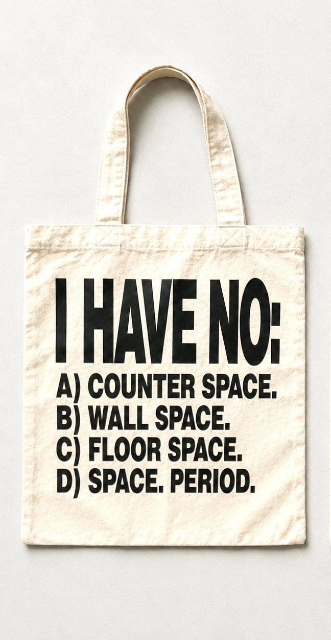 An off-white canvas tote bag with black printed text that reads 'I HAVE NO: A) COUNTER SPACE. B) WALL SPACE.